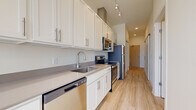 This single-floor unit showcases a thoughtfully designed interior with consistent light wood flooring running throughout the space. The galley-style kitchen is a standout feature, equipped with white shaker cabinetry extending to the ceiling, brushed nickel hardware, neutral quartz countertops, a stainless steel undermount sink, and a full suite of stainless steel appliances including a dishwasher, electric range, over-the-range microwave, and refrigerator. Track lighting overhead enhances functionality in the cooking area.

The living area benefits from floor-to-ceiling windows that flood the space with natural light and provide expansive views of the surrounding urban skyline, creating an open and airy atmosphere. The bathroom continues the clean aesthetic with white vanity cabinetry, a large mirror, light tile flooring, and a neutral color palette that maintains visual continuity with the rest of the unit.

Practical amenities include a dedicated in-unit laundry closet housing a stacked washer and dryer, as well as a walk-in closet with built-in shelving for organized storage. A barn-style sliding door adds a subtle modern design detail. The overall palette of whites, light wood tones, and soft neutrals creates a cohesive, contemporary environment throughout.