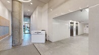 The Alexander's lobby and multi-purpose room at 5121 Bishop St in Halifax, NS, presents a generously proportioned ground-floor amenity space designed with a contemporary aesthetic. The entrance lobby features large-format tile flooring, exposed concrete columns, and soaring ceilings that create an immediate sense of openness and architectural character. A concierge desk welcomes residents upon arrival, flanked by a bank of mailboxes and elevator access.

Transitioning into the lounge area, the space opens dramatically with floor-to-ceiling glazing that frames views of the surrounding streetscape and fills the interior with abundant natural light. Exposed concrete structural columns anchor the room while complementing the clean, modern palette of white walls and neutral flooring. Seating arrangements include leather sofas and accent chairs in both dark and warm tan tones, centered around glass coffee tables and area rugs. A built-in fireplace with white brick surround and integrated shelving serves as a refined focal point.

The multi-purpose room is equipped with a long quartz-topped counter, slate-blue cabinetry, white tile backsplash, and circular pendant lighting, offering a functional and attractive space for events or resident use. A wall-mounted display screen adds versatility to the room. Throughout, the design balances modern sophistication with comfortable, inviting details suited to a vibrant urban community.