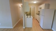 This single-floor space features a compact, efficiently arranged kitchen as its central highlight. The kitchen is equipped with white appliances, including a freestanding electric coil range, a full-sized refrigerator, and a sink set within white cabinetry that provides practical storage. An overhead cabinet is mounted above the range area, maximizing vertical storage space. The light hardwood-style flooring runs consistently throughout, lending warmth and visual continuity to the interior. Neutral beige walls complement the clean, understated aesthetic of the space. A hallway extends from the kitchen area, connecting to additional rooms accessed through white-framed doors, suggesting a straightforward and functional layout. Flush-mount ceiling lighting provides even illumination throughout. The overall ambiance is modest and practical, with a clean, move-in-ready feel characterized by its light tones, simple finishes, and efficient use of space.