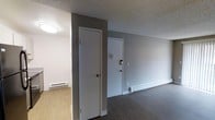 This single-floor 1-bedroom, 1-bathroom unit offers a straightforward, functional layout suited for everyday living. The entryway is visible from the main living area, featuring a solid entry door with a keypad lock for added security. Adjacent to the entry is a closet door, providing convenient storage near the front of the unit. The walls are finished in a neutral grey tone, complementing the dark laminate or vinyl plank flooring that runs through the main living space. A lighter-toned flooring is visible in a secondary area, likely the bedroom or kitchen, offering a subtle contrast. The ceilings feature a textured popcorn finish and are outfitted with flush-mount lighting. Baseboard heaters line the walls, serving as the primary heat source throughout the unit. White trim and baseboards provide a clean contrast against the grey walls and dark floors. The overall ambiance is minimal and neutral, creating a blank canvas that is easy to personalize. The unit's compact yet efficient design makes good use of the available space, with practical finishes that are durable and low-maintenance.