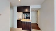 This single-floor space showcases a thoughtfully designed kitchen as its focal point, finished with rich dark espresso cabinetry that extends from floor to ceiling, creating a bold visual contrast against the white walls. The kitchen is equipped with stainless steel appliances, including a refrigerator, stove, and built-in oven, providing a fully functional cooking environment. A curved peninsula with a light granite countertop integrates a single-basin sink, offering both additional prep space and a natural boundary between the kitchen and the adjoining living area. Recessed lighting in the kitchen ensures the workspace is well-illuminated. The flooring transitions from light carpet in the living area to hardwood in the hallway beyond, adding warmth and definition between zones. A wide entryway corridor leads to a paneled white door, likely the main entrance, flanked by a closet with sliding doors — offering practical storage. The overall layout is efficient and cohesive, with a neutral color palette and modern finishes that give the space a clean, contemporary feel.