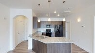This single-floor residence offers a thoughtfully designed open-concept layout that maximizes both functionality and style. The kitchen serves as the heart of the space, showcasing a spacious wraparound granite-topped island with an undermount sink, providing ample prep space and serving as a natural gathering point. Rich, dark wood cabinetry lines the walls from counter to ceiling, offering generous storage while contributing a sleek, contemporary aesthetic. Black appliances — including a range, built-in microwave, and a French door refrigerator — complement the darker tones of the cabinetry for a cohesive look. Three teardrop pendant lights hang above the island, casting warm, focused illumination that enhances the kitchen's inviting atmosphere. Recessed lighting further brightens the space, ensuring excellent visibility throughout. The kitchen flows seamlessly into the adjacent living areas via an arched architectural detail, adding a subtle traditional touch to the otherwise modern design. Light-toned wood-look flooring runs throughout, grounding the space and providing a clean contrast to the darker cabinetry. The overall ambiance is modern and functional, with a neutral color palette that offers a versatile backdrop for a variety of interior styles.