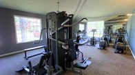 This fitness center offers a practical and well-appointed space designed to support a comprehensive workout routine. The room features a vaulted ceiling with an angled roofline that adds a sense of openness, complemented by a ceiling fan that promotes air circulation during exercise. Gray-painted walls provide a neutral, focused atmosphere, while durable flooring suitable for gym use runs throughout the space. Multiple windows along the perimeter flood the room with natural light and offer views of the surrounding outdoor environment, creating an inviting and energizing workout setting. The strength training area is anchored by a multi-station cable machine from Inflight Fitness, capable of supporting a wide variety of resistance exercises targeting multiple muscle groups. The cardio section is equipped with treadmills and an elliptical trainer, allowing users to engage in sustained aerobic exercise. The layout is efficiently organized, maximizing the available floor space to accommodate both strength and cardio equipment without feeling overcrowded. Overall, this fitness center provides a bright, functional, and well-maintained environment suitable for a range of fitness goals.