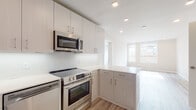 This thoughtfully designed single-floor unit showcases a clean, modern aesthetic with light-toned finishes and an open, airy feel throughout. The kitchen is a standout feature, outfitted with full stainless steel appliances including a refrigerator, range, dishwasher, and over-the-range microwave. Light wood-grain cabinetry extends to the ceiling, providing generous storage, while a textured penny-tile backsplash and crisp white countertops add refined detail. A peninsula counter enhances the layout, offering additional prep space and an open connection to the living area. Light hardwood-style flooring flows seamlessly throughout, reflecting natural light from large windows that frame urban views of the surrounding cityscape. The living areas are bright and open, enhanced by recessed lighting and neutral wall tones that create a welcoming, versatile environment. A dedicated closet houses a stacked washer and dryer unit, offering the convenience of in-unit laundry. The bathroom is finished with large-format light tile flooring, a floating vanity with under-cabinet lighting, a backlit mirror, and a full tub-shower combination. The overall design is cohesive and contemporary, emphasizing clean lines, quality materials, and functional living across every area of the space.