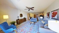 This single-floor residence offers an open-concept layout that seamlessly connects the living, dining, and kitchen areas. The living room serves as the central gathering space, anchored by a bold geometric-patterned area rug in blue, red, and yellow tones that adds a vibrant focal point to the room. Comfortable seating, including a tufted blue sofa and coordinating accent chairs, is arranged around a sleek glass coffee table, creating a relaxed yet stylish atmosphere. A wooden credenza with a round decorative mirror above it lines one wall, providing both storage and visual interest.

A ceiling fan with wooden blades is centrally mounted overhead, contributing to both comfort and airflow. The neutral-toned walls and light carpet flooring keep the space feeling bright and airy. Track lighting in the background illuminates the kitchen area, which features dark cabinetry, a breakfast bar with seating, and modern appliances. A doorway visible from the living space leads further into the unit, suggesting additional private rooms beyond. The overall design balances casual comfort with contemporary style.