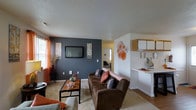 This single-story residence features four bedrooms and four bathrooms within a thoughtfully arranged floor plan. The living room serves as the central gathering space, highlighted by a bold slate-blue accent wall that anchors the room and provides a striking contrast to the surrounding white walls. A wall-mounted flat-screen television is centered on this feature wall, flanked by decorative artwork. The space is carpeted and receives natural light through a window dressed with warm orange curtains, adding a pop of color to the room. Decorative wall accents in coordinating tones contribute to the curated aesthetic. Adjacent to the living area, the kitchen is partially visible and features white cabinetry, a light countertop, and a pass-through or breakfast bar-style counter that connects the two spaces. The textured ceiling adds a classic residential touch throughout. The open sightlines between the living and kitchen areas create a connected, functional layout suitable for everyday living. With four bedrooms and four bathrooms, this residence offers ample accommodation and privacy for multiple occupants.