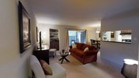 This single-floor residence offers a thoughtfully designed open-concept layout that seamlessly connects the living, dining, and kitchen areas. The living room serves as the heart of the home, centered around plush wall-to-wall carpeting and anchored by a comfortable seating arrangement. A built-in shelving unit with cabinetry provides both storage and display space, adding a refined architectural detail to the room. Neutral-toned walls keep the space feeling bright and inviting, while warm ambient lighting enhances the cozy atmosphere. A large sliding glass door floods the living area with natural light and opens directly to a private outdoor patio, offering a seamless indoor-outdoor connection. Adjacent to the living room, the dining area is defined by an elegant pendant light fixture that draws the eye upward and creates an intimate setting for meals. A pass-through or partial opening connects the dining space to the kitchen, maintaining an airy, communicative flow throughout the home. Together, the spaces strike a balance between comfort and functionality, making this a welcoming and practical single-level residence.