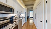 This efficiently designed one-bedroom, one-bathroom unit makes excellent use of its single-floor layout, offering a cohesive blend of modern aesthetics and practical functionality throughout. The galley-style kitchen is a standout feature, outfitted with white shaker cabinetry accented by matte black hardware, a classic subway tile backsplash, dark quartz countertops, and a full suite of stainless steel appliances. Warm wood-look flooring and subtle recessed lighting with tray ceiling details lend the kitchen an elevated, polished character. A sleek entry door with frosted glass sidelights introduces natural light at the front of the unit. The generously proportioned bedroom serves as a true retreat, boasting soaring ceilings with exposed beam details, warm hardwood flooring, and a set of full-height French doors that flood the room with natural light while offering outdoor access. The bathroom is finished to a high standard, featuring large-format gray floor tiles, a glass-enclosed walk-in shower with white subway tile surround, a white vanity with dark quartz countertop, and matte black fixtures throughout. A dedicated closet is conveniently accessible directly from the bathroom, completing this well-appointed space.