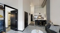 This contemporary single-floor loft showcases an industrial-modern aesthetic that is both striking and functional. Soaring ceilings draw the eye upward, creating an expansive, airy atmosphere throughout the open-concept living space. Polished concrete floors run seamlessly underfoot, adding an industrial edge that complements the overall design language.

The kitchen is a standout feature, boasting crisp white cabinetry paired with a dramatic dark island topped with a light countertop. A row of black-and-gold bar stools lines the island, making it an ideal spot for casual dining or entertaining. Stainless steel appliances, including a refrigerator and microwave, are neatly integrated into the cabinetry, while pendant lighting hangs above the island to provide focused illumination and a touch of modern elegance.

Bold black interior doors with clean panel detailing create strong visual contrast against the bright white walls, reinforcing the space's monochromatic design theme. Exposed ceiling infrastructure adds an authentic loft character, enhancing the sense of height and openness. The living area provides a welcoming transition from the kitchen, anchored by a patterned area rug and a marble-top accent table. Overall, this space delivers a refined yet edgy urban living experience.