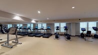 This fitness center offers a comprehensive workout environment designed to accommodate a wide range of exercise routines. The space is laid out across a single open floor, finished with durable speckled rubber flooring that provides both traction and comfort underfoot. Recessed lighting throughout the facility ensures the space is well-lit and inviting.

The cardio zone is lined with multiple treadmills and upright elliptical machines, each equipped with individual digital display screens. Large windows along the wall allow natural light to filter in, and full-length mirrors run along one side, visually expanding the room and allowing users to monitor their form.

The strength training area features a multi-tier dumbbell rack with a broad range of free weights, adjustable benches, and a cable pulley system for versatile resistance training. A dedicated functional training section includes a cable cross machine, a stability ball rack stocked with exercise balls in various sizes, and kettlebells.

The layout maintains generous open floor space at the center, enabling stretching, bodyweight exercises, and dynamic movements. The facility is accented by neutral wall tones and a clean, modern aesthetic throughout, contributing to a focused and professional training atmosphere. Glass-paneled entry doors complete the polished look of this well-appointed fitness space.