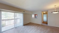 This single-floor residence offers a clean, functional layout designed for comfortable everyday living. The spacious living area is finished with wall-to-wall carpeting in a neutral tone, creating a warm and inviting atmosphere. Large vertical blinds frame a full-width sliding glass door that opens to an outdoor balcony, allowing abundant natural light to fill the room and providing a seamless connection to the outdoors.

A distinctive pass-through window connects the living area directly to the kitchen, enhancing the open feel of the space and allowing for easy interaction between rooms. The kitchen features warm wood-toned cabinetry and includes built-in appliances such as a microwave, maintaining a practical and tidy appearance.

A hallway visible from the living area leads further into the residence, suggesting additional rooms beyond, including what appears to be a carpeted bedroom area. The walls throughout are finished in a soft, neutral beige, contributing to the bright and airy character of the home.

Overall, the space presents a straightforward and well-maintained interior with consistent finishes, ample natural light, and a layout that balances open communal areas with private living spaces.