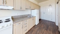 This single-floor unit offers a straightforward, functional layout designed with practicality in mind. The open-concept living and kitchen area is one of the space's defining features, where rich, dark wood-look flooring flows seamlessly from the kitchen into the adjacent living zone. A pendant light fixture hangs centrally, adding a touch of warmth to the area, while large windows with vertical blinds allow generous natural light to fill the room. The carpeted living area provides a comfortable contrast to the hard flooring surfaces.

The kitchen is efficiently designed, featuring cream-toned shaker-style cabinetry with brushed silver hardware extending both above and below the granite-patterned countertops. A stainless steel sink, white electric range, and full-size white refrigerator complete the kitchen's functional setup, with ample cabinet storage for everyday use.

The bathroom continues the wood-look flooring and is finished with clean white walls. It includes a wall-mounted sink, a mirrored medicine cabinet illuminated by a chrome light bar, and grab bars — indicating accessibility-conscious design. A towel bar and standard toilet round out the space, keeping the bathroom simple and easy to navigate.