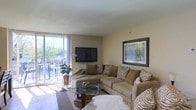 This single-floor space offers a welcoming and thoughtfully arranged living environment. The main living area is the focal point of the home, designed to maximize comfort and natural light. Large sliding glass doors span nearly the full width of the room, flooding the interior with abundant daylight and providing direct access to a private balcony that overlooks lush greenery — creating a seamless connection between the indoors and outdoors.

The living room is generously proportioned, accommodating a large L-shaped sectional sofa dressed in warm earth tones and complementary animal-print accent pillows, lending the space a cozy yet stylish character. A sleek wall-mounted flat-screen television serves as a practical centerpiece on one wall, while a framed decorative mirror adds visual depth and dimension to the adjacent wall. An oval glass-top coffee table anchors the seating arrangement, resting atop a neutral area rug that defines the lounging zone.

Soft, neutral-toned walls throughout contribute to an airy and calm ambiance, while vertical blinds on the sliding doors allow for adjustable light control and privacy. The overall atmosphere is inviting and livable, balancing functional design with warm, homey aesthetics — making this space well-suited for both relaxing and entertaining.