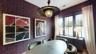 This intimate, single-floor room is defined by its bold, dramatic design aesthetic. Deep burgundy and gold patterned wallpaper envelops all four walls, establishing a rich, moody atmosphere throughout the space. A central oval table in a light wood tone serves as the focal point, surrounded by plush green velvet upholstered chairs that provide a striking color contrast against the dark walls.

Overhead, an industrial-style saucer pendant light with an amber glass globe fixture adds a sculptural, decorative element while providing warm ambient illumination. Recessed ceiling lights supplement the lighting scheme. A wall-mounted flat-screen television is positioned on one wall, indicating the room's function as a private meeting or conference space.

A large, dark-framed window along one wall allows natural light to filter in through sheer white curtains, with a low credenza beneath it holding a pair of matching table lamps. Two framed abstract artworks hang on adjacent walls, reinforcing the curated, design-forward character of the room.

A glass sliding partition wall connects this space to a larger, brighter adjoining lounge or common area, offering flexibility for private or open configurations. A patterned area rug anchors the seating arrangement, completing the cohesive and intentional interior design.