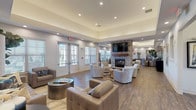 This single-story community clubhouse is thoughtfully designed to provide a welcoming and functional gathering space. The main lounge area serves as the centerpiece, featuring warm hardwood-style flooring, tray ceilings with recessed lighting, and multiple seating arrangements with neutral-toned sofas and patterned accent chairs. A dramatic floor-to-ceiling stone fireplace with a wood mantel anchors the room, complemented by a wall-mounted television above. Large French doors and shuttered windows allow abundant natural light to fill the space, creating a bright and inviting atmosphere.

Adjacent to the lounge, a dining area offers flexible seating around a round table, accented by striking metallic wall sculptures and contemporary artwork. A kitchenette nearby is equipped with granite countertops, dark cabinetry, a coffee station, and a wall-mounted television, making it well-suited for events and casual gatherings.

The fitness center features light teal walls, durable carpet tile flooring, floor-to-ceiling mirrors, and a comprehensive selection of equipment including treadmills, ellipticals, and a full dumbbell rack. A private restroom is accessible directly from the fitness area. Together, these spaces deliver a polished, amenity-rich environment designed for community engagement and everyday convenience.