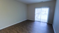 This single-floor space presents a clean, straightforward layout designed for comfortable living. The main living area is finished with rich, dark hardwood-style flooring that contrasts neatly against the neutral beige walls, creating a warm yet understated aesthetic. White baseboards frame the perimeter of the room, adding a crisp, finished appearance throughout.

A prominent sliding glass door fitted with vertical blinds serves as a key architectural feature, drawing in generous amounts of natural light and offering a view of the outdoors. The door provides both practical access to the exterior and a bright, airy quality to the interior space.

The ceiling incorporates a centrally placed HVAC vent, indicating a forced-air climate control system. The walls are smooth and free of ornate detailing, lending the space a modern, minimalist character that offers flexibility for a variety of furniture arrangements and décor styles.

Overall, the space conveys a functional and well-maintained environment. The combination of dark flooring, light-toned walls, and abundant natural light from the sliding door creates a balanced and inviting atmosphere suitable for everyday living.