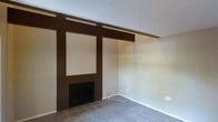 This two-floor space presents a straightforward yet characterful layout with notable design elements rooted in a mid-century aesthetic. The main living area is anchored by a fireplace set within a bold dark wood accent wall, which features structured paneling and a recessed display niche above the firebox. Horizontal wood beam detailing extends across the upper portion of the accent wall, adding a sense of architectural definition to the room. The flooring transitions from tile in the immediate fireplace area to carpet across the remainder of the living space, providing a practical and comfortable surface underfoot. Neutral beige walls keep the space feeling open and adaptable, while a textured popcorn ceiling adds a period-appropriate finishing detail. The living area offers ample wall space, lending flexibility for furniture arrangement. The two-level configuration of the overall property provides a natural separation of living and private spaces, a practical feature that adds functional depth to the home. Overall, the space carries a utilitarian character with vintage architectural touches that offer a solid foundation for personalization.