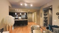 This well-appointed single-floor space showcases a thoughtfully designed open-concept layout that seamlessly connects the kitchen, dining, and living areas. The kitchen is a standout feature, boasting dark espresso cabinetry paired with granite countertops and a matching island with bar seating — ideal for casual dining or entertaining. Stainless steel appliances, including a full-size refrigerator and built-in microwave, add a modern, functional touch. Pendant lighting above the island and track lighting along the ceiling provide layered illumination throughout the space. Warm hardwood flooring extends across the main living areas, contributing to an inviting and cohesive aesthetic. Adjacent to the kitchen, the living area is accented by a built-in shelving unit, offering both display and storage space. A hallway leads to additional rooms, flanked by louvered doors that add a classic design element. The neutral color palette of soft grays and warm taupes creates a calm, contemporary ambiance throughout. Overall, the space combines practical design with modern style, making it both a comfortable and visually appealing environment.