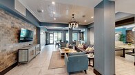 Located in Stuart, Florida, this single-floor community clubhouse offers a generously sized, thoughtfully designed gathering space that blends contemporary style with welcoming comfort. The main lounge area is anchored by a decorative pressed-tin ceiling and an industrial-style chandelier, creating a striking focal point overhead. Teal-toned walls, large-format tile flooring, and rich dark wood baseboards contribute to a cohesive, upscale aesthetic throughout. Comfortable seating arrangements — including tufted leather sofas and teal accent chairs — invite relaxed socializing around a central coffee table. A dedicated media alcove features a large flat-screen display set into a rustic brick accent wall, providing a cozy retreat for entertainment. The dining area showcases a live-edge wood communal table paired with sleek black chairs, bathed in natural light streaming through floor-to-ceiling glass doors and windows. These openings frame direct views of the outdoor pool courtyard, lined with palm trees and lounge seating. A long, well-lit corridor connects the various areas, while built-in shelving and a service counter offer additional functional detail. The overall ambiance is modern and inviting, designed to serve as a true hub for community life.