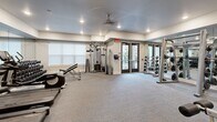 Located at 150 Country Squire Dr in Cromwell, Connecticut, this fitness center offers a thoughtfully designed exercise room that provides a comprehensive workout environment. The space features a clean, modern aesthetic with light gray walls, recessed lighting, and a ceiling fan that ensures comfortable air circulation throughout sessions.

The exercise room is well-appointed with a diverse selection of equipment to support a full-body workout routine. A row of Life Fitness treadmills with integrated display screens lines one side of the room, accompanied by additional cardio machines including a stair climber. On the strength training side, the space includes a multi-function cable machine, a leg extension/curl station, a dedicated free weight area with a full dumbbell rack, and a weight plate rack with barbell. A flat workout bench adds further versatility.

Floor-to-ceiling mirrors span an entire wall, amplifying the sense of space and allowing users to monitor their form during exercise. A wall-mounted flat-screen television provides entertainment during workouts. The room is finished with durable, cushioned carpet flooring suitable for fitness use, and a set of glass-paneled double doors allows natural light to filter in while providing easy access. Posted guidelines ensure a respectful, well-managed environment for all users.