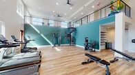 Located at 2950 Richmond Row Drive in Suwanee, Georgia, this two-story fitness center is a thoughtfully designed facility that combines functionality with striking architectural character. The main floor makes an immediate impression with soaring double-height ceilings, bold teal accent walls, and light hardwood flooring that runs throughout both levels. Recessed lighting and ceiling fans maintain a bright, comfortable atmosphere, while large windows allow natural light to pour in, offering views of the surrounding community.

The ground floor is equipped with a comprehensive selection of strength and resistance machines, adjustable benches, a dumbbell rack, and cable systems, providing a well-rounded space for resistance training. A dramatic wrought-iron spiral staircase serves as both a functional connection between floors and a visual centerpiece, leading up to the open mezzanine level above.

The upper level overlooks the main floor through floor-to-ceiling glass panels and metal railings, creating a sense of openness and visual continuity. This second floor is dedicated to cardio training, housing water rowing machines, stationary cycling bikes, and treadmills positioned beside windows with outdoor views. Additional accessories such as stability balls and resistance equipment are also available on this level, rounding out the facility's comprehensive fitness offering.