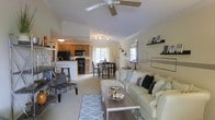 This single-floor residence showcases an open-concept layout that effortlessly connects the main living areas, creating a cohesive and functional flow throughout. The living room serves as the heart of the home, featuring plush neutral-toned seating, a wood-and-metal coffee table, and a decorative geometric-patterned area rug. A ceiling fan with a light fixture overhead ensures year-round comfort, while crown molding lends a refined architectural touch to the space. An accent wall with decorative shelving and a framed mirror adds visual interest and personality to the room. Adjacent to the living area, a cozy dining space accommodates a dark-wood table and chairs, positioned near a window that invites natural light into the home. The kitchen is well-appointed with warm wood cabinetry, black appliances including a range, microwave, and refrigerator, and a breakfast bar with seating for casual dining. Light-toned carpeting runs through the main living and dining areas, complementing the warm, neutral color palette that unifies the entire space. Overall, the home presents a welcoming and well-organized interior that balances comfort with tasteful design details.