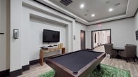 This single-floor game room is designed with both style and function in mind, offering a well-appointed space ideal for entertainment and leisure. The centerpiece of the room is a full-size pool table with a dark felt surface, positioned atop a rich green area rug that contrasts neatly with the light wood-look tile flooring running throughout the space. Recessed lighting set within a tray ceiling provides even illumination, while dark baseboards and door trim add a sharp, defined contrast against the neutral white walls.

A wall-mounted flat-screen television is positioned within a recessed architectural niche, accompanied by a wooden console table below. One wall features a bold vertical gold-and-white striped accent, adorned with decorative starburst wall art, adding visual interest and a touch of glamour. A triptych of large black-and-white equestrian canvas prints lines an adjacent wall, lending an artistic, thematic quality to the space.

The room opens through an arched corridor into an adjoining hallway, suggesting connectivity to broader amenity areas. Dark-framed doors and clean architectural lines throughout reinforce the space's polished, contemporary aesthetic, making it a well-designed venue for recreation.