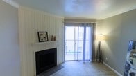 This single-floor space offers a welcoming and functional layout designed for comfortable everyday living. The living room serves as the heart of the home, anchored by a charming white-painted fireplace set against vertical shiplap-style wall paneling, adding a classic and cozy character to the room. Soft, neutral-toned carpet runs throughout, complementing the warm, understated color palette of the walls and trim. Crown molding lines the ceiling, adding a refined architectural touch that elevates the overall aesthetic. A textured ceiling adds subtle visual interest overhead. Natural light fills the living area through a large sliding glass door dressed with sheer vertical blinds, which open to a private balcony enclosed by a metal railing — offering a pleasant outdoor extension of the living space and a glimpse of the surrounding environment. The overall ambiance of the space is calm and inviting, with classic design elements that provide a neutral canvas well-suited for a variety of interior styles. The single-floor layout ensures easy accessibility and a straightforward, cohesive flow throughout the home.