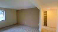 This single-floor space offers a clean and functional layout with a notably distinctive architectural feature: a vaulted ceiling that adds a sense of openness and volume to the main room. The angled ceiling line creates a dynamic visual element, drawing the eye upward and contributing to an airy, spacious atmosphere. The room is finished with wall-to-wall carpet in a neutral gray tone, providing a soft and comfortable underfoot experience that complements the warm, taupe-colored walls. The overall color palette is understated and cohesive, lending the space a calm, move-in-ready aesthetic. Adjacent to the main room, a walk-in closet offers practical storage with built-in shelving units visible through the open doorway. The shelving spans the full height of the closet space, maximizing vertical storage capacity. The transition between the main room and the closet is framed by a clean white doorframe, maintaining a consistent and tidy finish throughout. Electrical outlets are positioned along the baseboards, and the walls are smooth and well-maintained. The space is well-suited for use as a bedroom, offering both comfort and storage in a straightforward, uncluttered layout.