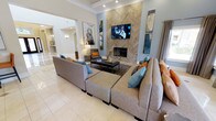 This single-floor space centers around a well-appointed living area that immediately draws the eye to its defining architectural feature: a floor-to-ceiling herringbone wood-panel accent wall housing both a sleek fireplace and a wall-mounted flat-screen television. The tall ceilings amplify the sense of openness, while recessed lighting and crown molding add refined detail overhead. Light neutral tile flooring extends throughout, contributing to the bright, airy atmosphere. The living area is furnished with a generous sectional sofa in soft gray, accented with teal and burnt orange throw pillows that introduce color and warmth. A rectangular upholstered ottoman serves as a central coffee table, anchoring the seating arrangement over a patterned area rug. Flanking the fireplace wall, vertical art panels complement the space's cool blue-gray color palette. An open archway leads to an adjoining hallway or secondary area, reinforcing the flowing, connected layout of the home. The overall ambiance is modern yet inviting, blending clean architectural lines with thoughtfully curated furnishings and décor.