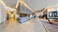 This single-floor space presents a compelling contrast between two distinct areas. The lobby is an architectural standout, showcasing a sweeping, floor-to-ceiling wood-paneled reception desk accented by warm indirect cove lighting that traces the curves of the ceiling. Polished concrete floors extend throughout the expansive entrance, lending a clean, contemporary feel. The open layout allows for generous circulation space, while a lounge area in the background features neutral-toned seating amid indoor greenery, contributing to a welcoming and professional ambiance. Vertical slatted screens further define zones within the lobby without compromising its sense of openness.

The bathroom offers a practical and neatly appointed layout. It includes a vanity with a white countertop sink, brushed nickel faucet, and a warm wood-toned cabinet below. A wood-framed mirror is mounted above the vanity, and white floral wall décor adds a subtle personal touch. Natural light filters in through a window fitted with pleated blinds, keeping the space bright. A shower with a floral curtain and an over-toilet shelving unit provide additional storage and functionality. The bathroom's white walls and trim create a clean, neutral backdrop throughout.