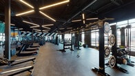 This contemporary fitness center is designed to deliver a comprehensive workout experience across a single, generously sized floor. The main gym floor features an extensive array of strength and conditioning equipment, including Matrix machines, free weights, and cable systems, all set beneath a dramatic dark ceiling fitted with bold linear LED lighting. Vibrant geometric and scenic murals adorn the walls, energizing the space with color and movement.

A dedicated cardio zone offers treadmills, ellipticals, stair climbers, and stationary bikes positioned directly in front of expansive floor-to-ceiling windows, providing sweeping city skyline views that make every workout engaging. An adjacent functional training area is equipped with TRX suspension systems, sled tracks, and multi-use rigs, bathed in natural light from large industrial-style windows.

A refined studio space features warm hardwood flooring, wall-to-wall mirrors with integrated LED backlit surrounds, wall-mounted ballet barres, and delicate floral murals — ideal for barre, yoga, or dance classes. A welcoming lounge area with stylish seating, polished concrete floors, and multi-tonal blue-painted walls provides a relaxed transition zone. Dual water fountains with a bottle-fill station and a glossy dark tile accent wall complete the thoughtfully designed amenity spaces throughout.