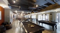 This clubroom is a thoughtfully designed social and recreational space that blends industrial aesthetics with comfortable, inviting elements. The open-plan layout accommodates multiple activity zones, anchored by two full-size billiard tables set on polished concrete floors. Exposed black ductwork and ceiling infrastructure lend an urban, warehouse-inspired character overhead, while pendant lighting with black metal shades provides focused illumination throughout the space. A warm contrast is introduced through brick accent walls, which add texture and visual interest alongside the darker industrial finishes. The lounge area features generous seating with sofas and chairs arranged to encourage conversation and relaxation. Toward the perimeter, bar-height tables with metal chairs offer additional seating options for dining or socializing. Large windows along the far wall flood the space with natural light and provide views to the outdoors, creating an airy feel that balances the bold, dark design elements. A kitchen or bar area is visible in the background, supporting catered events or casual use. A wall-mounted television adds to the entertainment options available. Overall, the clubroom delivers a dynamic, multi-functional environment well-suited for both casual hangouts and organized social events.