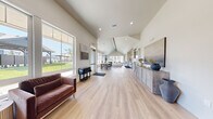 Located at 2700 Pierce Pkwy in Springfield, Oregon, this single-floor community clubhouse offers a thoughtfully designed, open-concept layout that balances functionality with modern aesthetic appeal. Light oak luxury vinyl plank flooring runs throughout, unifying the space and reflecting an abundance of natural light that pours in through oversized windows and glass doors.

The main gathering area features vaulted ceilings that amplify the sense of spaciousness, complemented by recessed lighting and warm-toned wall sconces. A built-in lounge zone along one wall incorporates bench seating with small accent tables, while leather armchairs and ottoman-style stools provide flexible social seating arrangements. A bold charcoal accent wall anchors this area with contrast and depth.

The kitchenette is well-appointed with light maple cabinetry, a white tile backsplash, stainless steel appliances including a full-size refrigerator and dishwasher, open display shelving, and a countertop workspace — ideal for community events and gatherings.

A dedicated dining area features a large rustic wood table with wishbone chairs, bathed in natural light from a trio of gridded windows overlooking a landscaped outdoor setting and community pavilion. An additional sun-drenched lounge nook at the far end offers direct views of the outdoor pool area. The entry foyer provides a welcoming transition with tasteful décor and a glass-panel exit door.