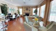 This thoughtfully designed single-floor space seamlessly blends comfort and functionality in an open, airy layout. The main living area centers around a elegant plastered fireplace adorned with a decorative sunburst mirror, serving as a striking focal point. Warm hardwood flooring flows throughout, complementing the neutral, earthy tones of the walls and furnishings.

The living area is furnished with plush, neutral-toned seating accented with teal and turquoise throw pillows, creating a fresh and inviting atmosphere. Adjacent to the seating area, a glass-top dining table surrounded by patterned upholstered chairs provides a refined space for gatherings, accented by a vibrant floral centerpiece.

Large windows dressed with warm-toned drape panels and slatted blinds allow generous natural light to fill the room while offering privacy, and offer views of lush greenery outside. Potted indoor plants strategically placed throughout add a natural, refreshing touch.

A dedicated workspace is also integrated into the room, featuring a desk, monitor, and white ergonomic chairs — making this space equally suited for productivity. Crown molding and recessed lighting add refined architectural detail, completing the polished yet welcoming character of the space.