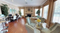 This thoughtfully designed single-floor space seamlessly blends comfort and functionality in an open, airy layout. The main living area centers around a elegant plastered fireplace adorned with a decorative sunburst mirror, serving as a striking focal point. Warm hardwood flooring flows throughout, complementing the neutral, earthy tones of the walls and furnishings.

The living area is furnished with plush, neutral-toned seating accented with teal and turquoise throw pillows, creating a fresh and inviting atmosphere. Adjacent to the seating area, a glass-top dining table surrounded by patterned upholstered chairs provides a refined space for gatherings, accented by a vibrant floral centerpiece.

Large windows dressed with warm-toned drape panels and slatted blinds allow generous natural light to fill the room while offering privacy, and offer views of lush greenery outside. Potted indoor plants strategically placed throughout add a natural, refreshing touch.

A dedicated workspace is also integrated into the room, featuring a desk, monitor, and white ergonomic chairs — making this space equally suited for productivity. Crown molding and recessed lighting add refined architectural detail, completing the polished yet welcoming character of the space.