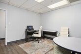 This professional business center office is a thoughtfully designed single-room workspace that balances focused individual work with collaborative functionality. The room features a drop-panel acoustic ceiling fitted with recessed fluorescent lighting that ensures consistent, even illumination throughout the space.

The interior is anchored by a clean, neutral color palette — crisp white walls on three sides contrast with a bold navy blue accent wall that adds depth and visual interest to the room. Light-toned wood-look plank flooring runs throughout, contributing a warm, grounded feel to the otherwise modern aesthetic, and is complemented by gray baseboards that provide a clean finish along the perimeter.

The layout is divided into two functional zones. A dedicated workstation area includes a white rectangular desk paired with a cushioned, high-back white office chair, suitable for computer work and individual tasks. A colorful area rug beneath the desk adds texture and defines this section of the space. On the opposite side, a small collaboration area features a round white table and an upholstered ottoman-style seat, ideal for brief meetings or consultations.

A wood-paneled door provides access to the broader office environment. Multiple electrical outlets are conveniently positioned along the walls, ensuring full connectivity for office equipment. The overall ambiance is clean, modern, and professional — well-suited for focused work or private client meetings.