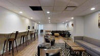 This contemporary lounge space is thoughtfully designed to accommodate a variety of social and gathering needs within a single open-plan layout. The space features a drop ceiling fitted with recessed pot lights that cast a warm, even illumination throughout, creating an inviting and comfortable atmosphere. Light hardwood-style flooring runs across the entire area, providing a clean and modern foundation that complements the neutral white walls.

The layout is organized into several distinct seating zones. A large L-shaped dark sectional sofa anchors one area, paired with a wooden coffee table and positioned atop a bold black-and-white diamond-patterned area rug. Nearby, a cluster of sleek modern swivel chairs with metallic bases surrounds glass-top accent tables, offering a more intimate conversational setting. A row of industrial-style bar stools lines a long counter along one wall, providing an additional casual seating option. Additional lounge seating is arranged throughout to maximize capacity and flexibility.

Decorative wall art, including metal sculptural pieces and a warm-toned canvas painting, add visual interest and personality to the space. The overall design strikes a balance between modern sophistication and relaxed functionality, making it well-suited for social gatherings, events, or casual everyday use.