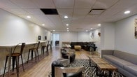 This contemporary lounge space is thoughtfully designed to accommodate a variety of social and gathering needs within a single open-plan layout. The space features a drop ceiling fitted with recessed pot lights that cast a warm, even illumination throughout, creating an inviting and comfortable atmosphere. Light hardwood-style flooring runs across the entire area, providing a clean and modern foundation that complements the neutral white walls.

The layout is organized into several distinct seating zones. A large L-shaped dark sectional sofa anchors one area, paired with a wooden coffee table and positioned atop a bold black-and-white diamond-patterned area rug. Nearby, a cluster of sleek modern swivel chairs with metallic bases surrounds glass-top accent tables, offering a more intimate conversational setting. A row of industrial-style bar stools lines a long counter along one wall, providing an additional casual seating option. Additional lounge seating is arranged throughout to maximize capacity and flexibility.

Decorative wall art, including metal sculptural pieces and a warm-toned canvas painting, add visual interest and personality to the space. The overall design strikes a balance between modern sophistication and relaxed functionality, making it well-suited for social gatherings, events, or casual everyday use.