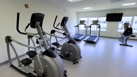 This single-level fitness room offers a focused cardio-oriented workout environment. The space is designed with simplicity and functionality in mind, featuring bright white walls and a smooth, light-colored flooring that together create a clean, clinical aesthetic. Natural light filters in through large windows along one wall, providing views of the surrounding exterior and contributing to an open, airy atmosphere.

The room is equipped with multiple elliptical cross-trainers positioned along one side, offering low-impact cardiovascular workout options. Opposite these, two treadmills are aligned facing the windows, allowing users to exercise while enjoying the natural light and outdoor views. The equipment appears modern and well-maintained, with digital display consoles on each machine.

The layout is straightforward, with sufficient space between machines to allow comfortable use without overcrowding. Overhead, a recessed ceiling light fixture supplements the natural light, ensuring the room remains well-illuminated at all times. The overall ambiance is utilitarian yet welcoming, providing an efficient and practical space suited for regular cardiovascular exercise.
