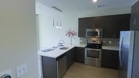 This single-floor space showcases a thoughtfully designed, contemporary kitchen that serves as a central highlight. The kitchen features dark espresso cabinetry paired with crisp white quartz countertops, creating a striking visual contrast. A full suite of stainless steel appliances — including an electric range, over-the-range microwave, and dishwasher — adds a polished, modern touch. A neutral-toned subway tile backsplash ties the design together with understated elegance. The peninsula layout provides both functional workspace and a casual dining or entertaining area, complete with pendant lighting overhead. Three sleek, tapered pendant lights hang above the peninsula, offering warm, focused illumination. Light wood-style flooring runs throughout, complementing the overall cool, neutral color palette and adding a sense of warmth to the space. The open layout allows for easy movement and a sense of spaciousness, while clean lines and minimalist finishes give the interior a refined, contemporary ambiance. The bright white walls enhance the feeling of openness and allow natural and artificial light to reflect evenly throughout the space.