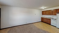 This single-floor unit offers a straightforward, functional layout designed for comfortable everyday living. The open living and dining area features neutral carpet flooring and a textured ceiling, with a window unit providing climate control and a window fitted with blinds that allows natural light to filter in. The space flows seamlessly into the kitchen, which is defined by warm medium-toned wood cabinetry, light tile flooring, and white appliances including an electric range, refrigerator, and a sink positioned beneath upper cabinets. A built-in counter with additional cabinetry provides supplementary storage at the kitchen's edge, reinforcing the practical design throughout.

The bedroom is a well-proportioned, carpeted room featuring a ceiling fan with integrated lighting and a window with blinds that brings in natural light. A closet with a rod is accessible nearby, offering organized storage. The bathroom is clean and utilitarian, featuring a stand-alone shower stall with a curtain rod and chrome fixtures, alongside a standard toilet. The textured walls throughout add subtle visual character to the otherwise neutral palette. Overall, the unit presents a clean, move-in-ready space with consistent finishes throughout.