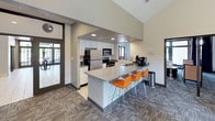 This thoughtfully designed single-floor space blends functionality with a welcoming, contemporary aesthetic. The kitchen area is a standout feature, boasting soaring vaulted ceilings, white cabinetry, stainless steel appliances including a refrigerator, range, and microwave, and a generous gray-topped peninsula with sleek orange bar stools that add a bold pop of color. A coffee station is conveniently positioned nearby, making it an ideal gathering spot for casual socializing or morning routines. The open layout transitions seamlessly into multiple lounge and seating areas, all finished with a coordinated patterned carpet in neutral tones. One lounge zone offers a cozy arrangement of blue upholstered chairs, colorful ottomans with southwestern-inspired patterns, and small accent tables, anchored by a vibrant large-format abstract painting on the wall. Another area provides a mix of small round café tables with orange metal chairs, creating a lively and informal atmosphere ideal for conversation or light work. Large glass doors and windows throughout the space invite abundant natural light and provide direct views and access to the surrounding greenery outdoors. The overall ambiance is modern, energetic, and inviting, combining thoughtful furnishings and curated artwork to create a comfortable and functional communal environment.