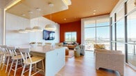 Located at 3615 Tryclan Drive in Charlotte, NC, this single-floor open-plan space seamlessly integrates a living room, dining area, and kitchen into one cohesive, thoughtfully designed layout. The interior makes an immediate visual impact with its bold terracotta orange accent walls and ceilings, which contrast strikingly against the crisp white surfaces throughout. Light hardwood flooring runs continuously across the entire space, lending warmth and a sense of continuity.

The kitchen and dining area feature a sleek white island with bar seating, illuminated by a row of modern pendant lights suspended from a structured ceiling detail. The living area is anchored by a wall-mounted flat-screen television and opens into a bright seating nook framed by dramatic floor-to-ceiling curtain-wall windows. These expansive glazed walls flood the space with natural light and offer panoramic views of the Charlotte skyline and surrounding neighborhood, making the outdoor scenery a defining architectural feature.

A second seating area near the glass wall provides an elevated vantage point, ideal for taking in the cityscape. Decorative wall art and natural wood accent pieces contribute to the warm, contemporary aesthetic. The open layout, abundant natural light, and seamless indoor-outdoor visual connection give this space a modern, sophisticated ambiance suited for both relaxed living and entertaining.