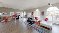 This single-floor clubhouse is a welcoming community hub designed with both functionality and style in mind. The space is immediately distinguished by its series of tall arched windows that line the walls, flooding the interior with generous natural light and offering views of the surrounding exterior. Vaulted ceilings add a sense of openness and airiness throughout.

Rich hardwood-style flooring flows seamlessly across the entire space, grounding the vibrant, contemporary décor. The main lounge area is a standout feature, furnished with a sleek white sectional sofa accented by bold magenta throw pillows, paired with modern black armchairs and glass-top accent tables. A striking multicolored area rug anchors the seating arrangement, adding an energetic burst of color that sets the tone for the entire space.

A dedicated leasing office area includes a large workstation, a round glass-top table with bright orange chairs, and colorful abstract artwork on the walls — creating a professional yet inviting environment. Toward the rear, French doors open to the exterior, while a service counter and entertaining elements such as a popcorn machine add a fun, community-centered character.

Throughout the clubhouse, vibrant abstract paintings adorn the walls, and lush potted greenery provides natural accents. Ceiling fans ensure comfortable airflow, completing this lively and well-appointed community gathering space.
