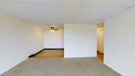 This single-floor space is defined by its clean, functional layout and practical design elements. The interior is divided by a partial wall, creating two distinct zones within an open floor plan. The carpeted section offers a softer, more comfortable surface underfoot, while the adjacent area transitions to smooth hardwood flooring, providing a contrast in texture and tone.

The drop tile ceiling runs throughout the space, giving it a uniform, finished appearance. A ceiling fan with an integrated light fixture is installed in the hardwood-floored section, providing both air circulation and ambient lighting. Dark baseboards line the perimeter of the walls, adding a subtle contrast to the neutral, white wall surfaces.

A hallway visible beyond the partial dividing wall leads to additional rooms, including a door that hints at further private spaces within the unit. Electrical outlets and light switches are thoughtfully placed throughout. The overall ambiance is neutral and versatile, offering a blank canvas suitable for a variety of uses and furnishing styles. The layout promotes a practical flow between areas while maintaining a sense of openness.