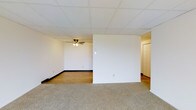This single-floor space is defined by its clean, functional layout and practical design elements. The interior is divided by a partial wall, creating two distinct zones within an open floor plan. The carpeted section offers a softer, more comfortable surface underfoot, while the adjacent area transitions to smooth hardwood flooring, providing a contrast in texture and tone.

The drop tile ceiling runs throughout the space, giving it a uniform, finished appearance. A ceiling fan with an integrated light fixture is installed in the hardwood-floored section, providing both air circulation and ambient lighting. Dark baseboards line the perimeter of the walls, adding a subtle contrast to the neutral, white wall surfaces.

A hallway visible beyond the partial dividing wall leads to additional rooms, including a door that hints at further private spaces within the unit. Electrical outlets and light switches are thoughtfully placed throughout. The overall ambiance is neutral and versatile, offering a blank canvas suitable for a variety of uses and furnishing styles. The layout promotes a practical flow between areas while maintaining a sense of openness.