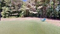 Located at 1000 Park Place Ave in Morrisville, North Carolina, this outdoor amenity space offers a well-appointed and inviting environment designed for relaxation and recreation. The space is framed by mature trees and dense natural greenery, creating a serene, park-like atmosphere that feels both private and open.

A standout feature is the synthetic putting green, providing a smooth, well-maintained surface ideal for casual golf practice. Nearby, a pergola structure serves as a covered gathering area, outfitted with comfortable outdoor seating and anchored by string lights that lend a warm, social ambiance during evening hours. Adjacent to the pergola, a concrete patio extends the usable outdoor space, accommodating additional seating arrangements for lounging or socializing.

The expansive open lawn area offers generous green space for a variety of outdoor activities, bordered by natural wooded edges that provide shade and a sense of seclusion. String lights are strung across poles throughout the space, adding a charming aesthetic touch that transitions seamlessly from day to night use. A utility structure with warm wood cladding is also present on-site, complementing the natural surroundings. The overall design balances functional recreational amenities with a relaxed, nature-integrated setting, making it an appealing outdoor destination.