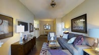 This single-floor residence offers an open-concept layout that seamlessly connects the living, dining, and kitchen areas, creating a spacious and cohesive flow throughout the home. The living room is anchored by a plush gray sofa accented with purple and teal throw pillows, complemented by a glass-top coffee table and a patterned area rug in neutral tones with purple detailing. Framed artwork adorns the walls, adding a curated, gallery-like feel to the space. A ceiling fan overhead and warm ambient lighting contribute to the comfortable atmosphere. The dining area sits just beyond the living space, featuring a compact dining table with chairs beneath an elegant pendant light fixture, with a decorative mirror mounted on the wall behind it. The kitchen, visible in the background, showcases white cabinetry and a breakfast bar that opens to the dining space, enhancing the open and airy character of the home. Dark hardwood-style flooring runs throughout, providing a rich contrast to the soft, neutral wall tones. The vaulted ceiling adds a sense of vertical spaciousness, making the entire living area feel bright, open, and inviting.