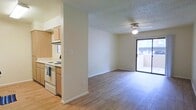 This single-floor space offers a clean, open layout that seamlessly connects the kitchen and living area, creating a sense of openness and easy flow throughout. The kitchen is positioned adjacent to the main living space, separated by a partial wall that maintains visual connectivity while defining each zone. It features light wood cabinetry, a white electric range, an overhead range hood, and a window that draws in natural light. The living area is generously proportioned, showcasing warm hardwood-style flooring that runs throughout and a ceiling fan that adds both function and a casual charm. A textured ceiling adds subtle architectural detail across the space. Neutral, light-toned walls contribute to a bright and welcoming ambiance. A large sliding glass door at the far end of the living area fills the room with natural light and provides direct access to an enclosed outdoor patio, offering a seamless transition between interior and exterior living. The overall design is practical and inviting, with clean lines and a versatile open floor plan well-suited for everyday living.